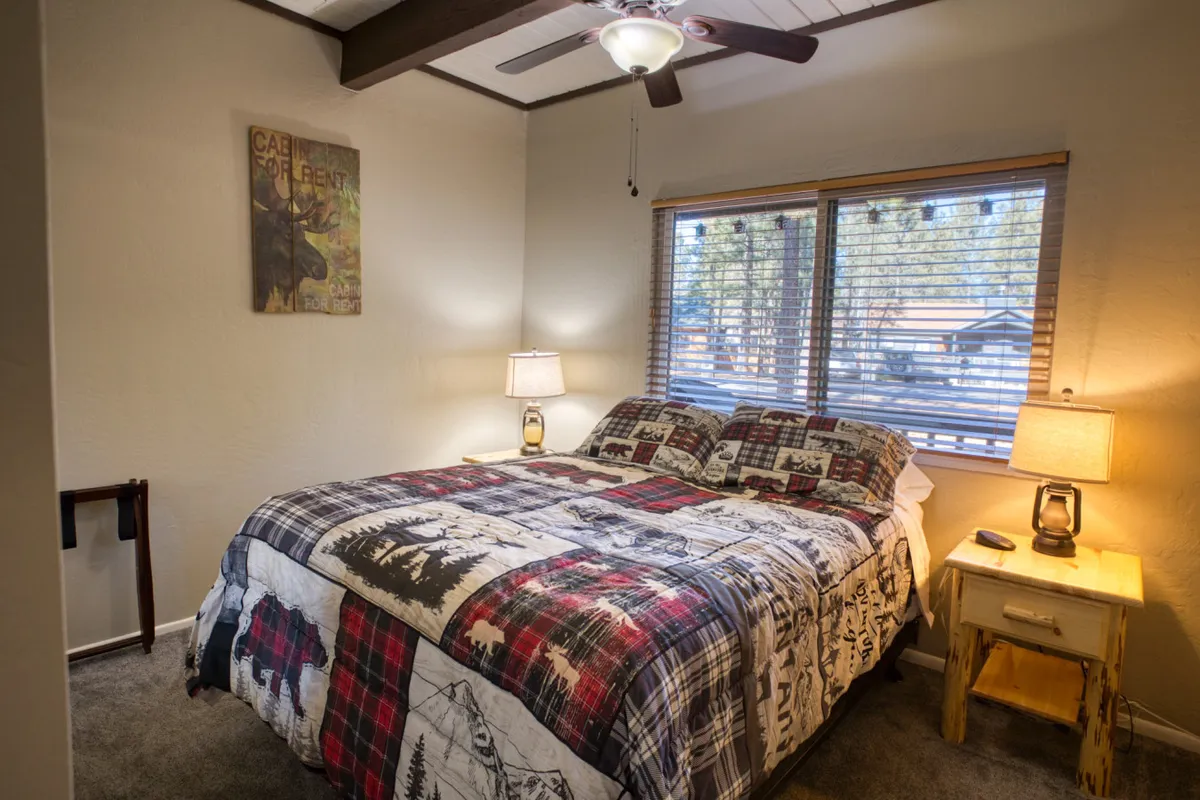 A cozy bedroom featuring a bed with a patterned quilt, flanked by wooden nightstands. Large windows with blinds fill the space with inviting natural light, creating an atmosphere perfect for relaxing or enjoying the morning sun.
