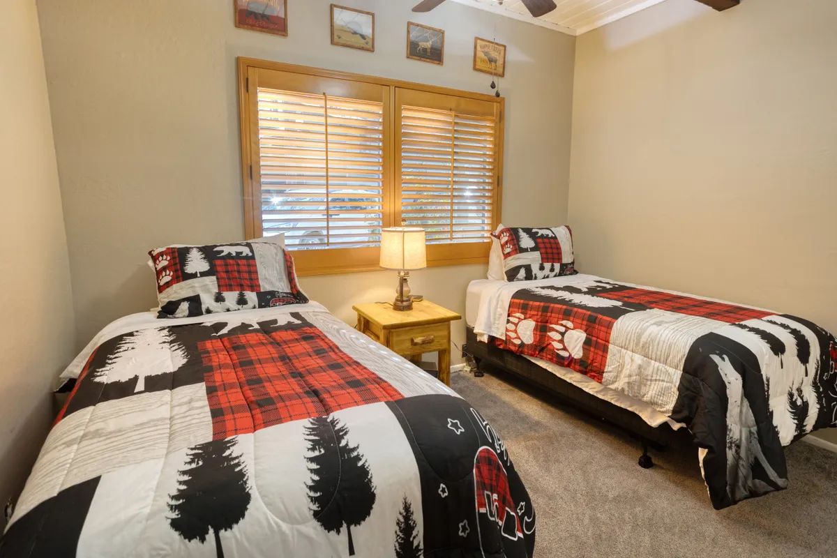 A cheerful guest room with twin beds, neatly dressed with coordinated linens, a bedside table with a reading lamp, a bright window with blinds, and decorative wall art. The welcoming ambiance suggests a perfect spot for kids or friends to unwind.