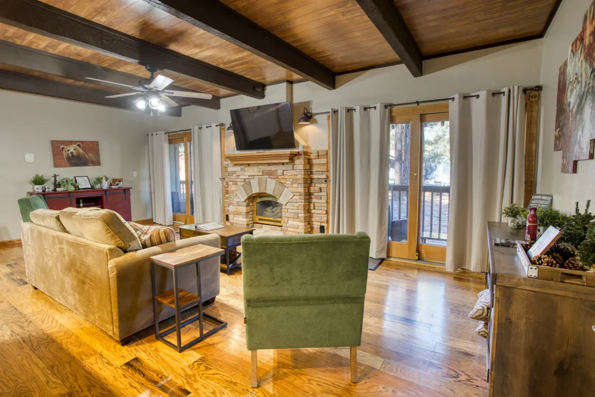 This image captures a warm, inviting living space with a cozy stone fireplace as the focal point. Plush seating, including a sofa and a green chair, are arranged for relaxation and conversation. The wooden floor adds rustic charm, while ample natural light streams through large windows, subtly connecting the indoors to the beauty outside. A TV is present, suggesting the space is ideal for both quiet evenings and lively gatherings.