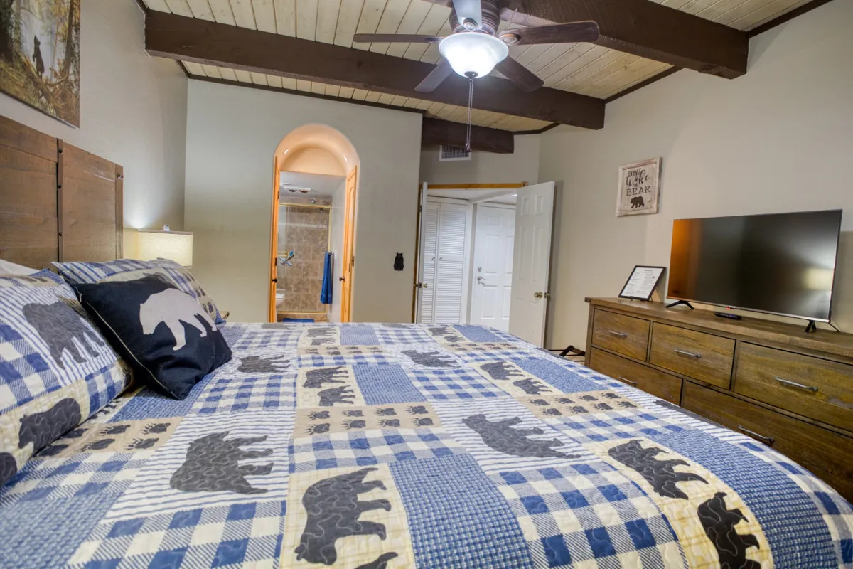 A spacious bedroom featuring a large bed with patterned bedding, complemented by wooden furniture, a convenient wall-mounted television, and a ceiling fan overhead. The space is bathed in soft natural light, inviting relaxation and comfort.